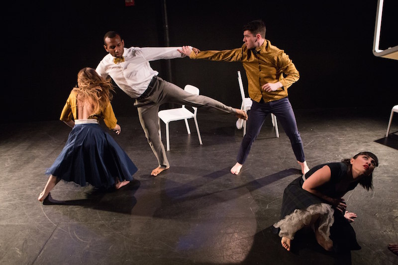 One dancer looks up as she crouches downstage left. One male dancer holds another by his wrists. A woman her back to the audience revealing a backless blouse.